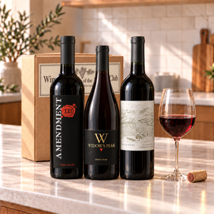 Illustrative three-bottle red wine selection on marble with Wine of the Month Club branded shipping box behind the bottles